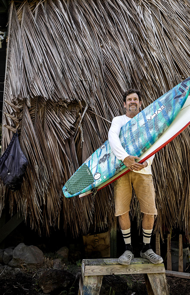 Man stands in front of a hut roof made from dried palm tree branches holding his blue and white surf board