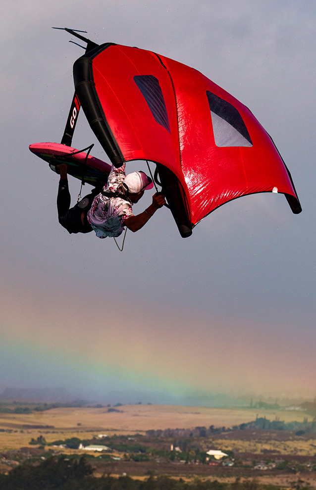 A person glides thru the air after launching from the water using a watercraft and surf board. In the background shows clear skies and a rainbow