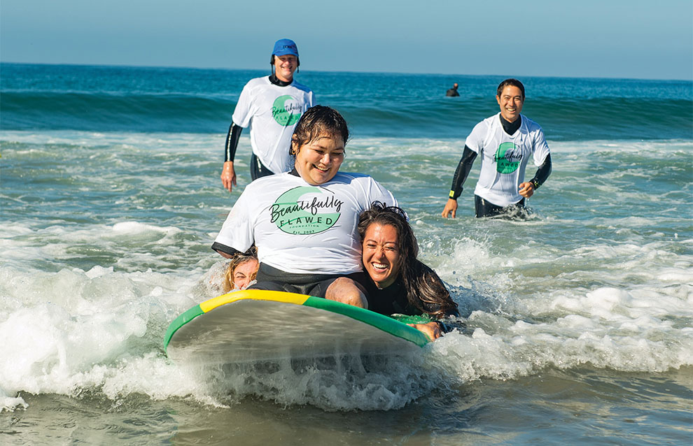 girl with disability rides surfboard toward shore with support from surf coach.