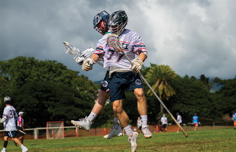 two men's lacrosse players jump in air together during a game on the field.