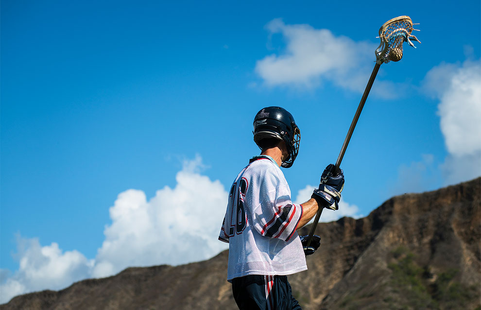 one lacrosse player with jersey and helmet holds lacrosse stick against blue skies and mountains in the back.