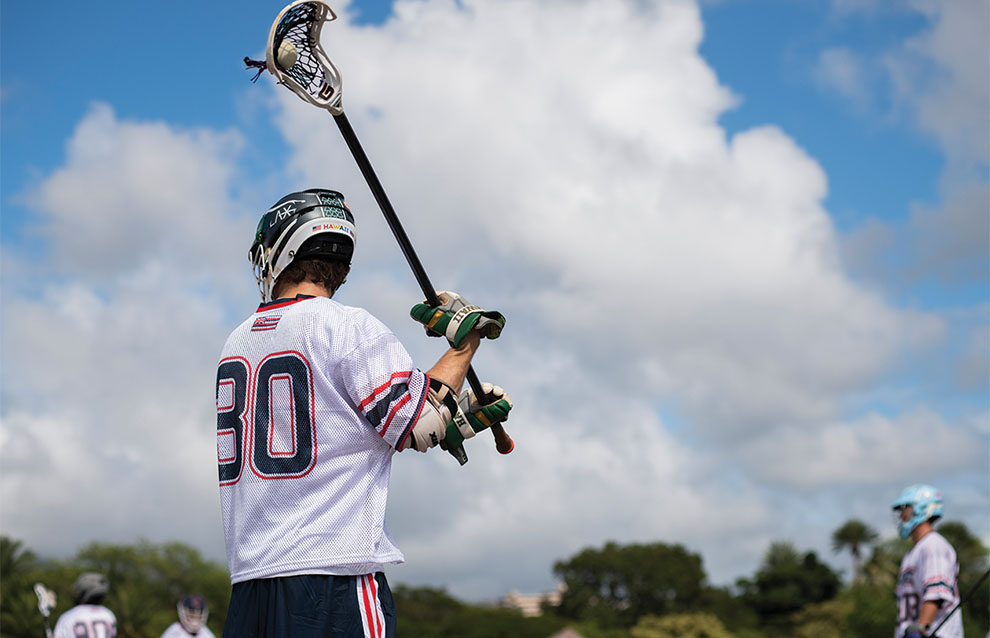 lacrosse player with white jersey on the field holds up stick.