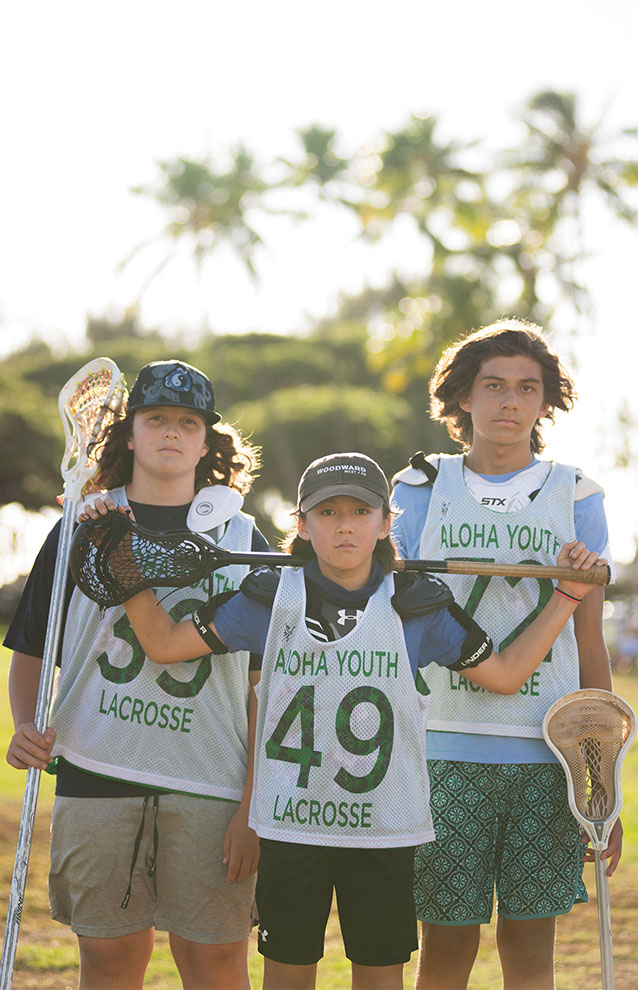 three boys stand together with lacrosse jerseys while shortest boy in front holds lacrosse stick behind neck.