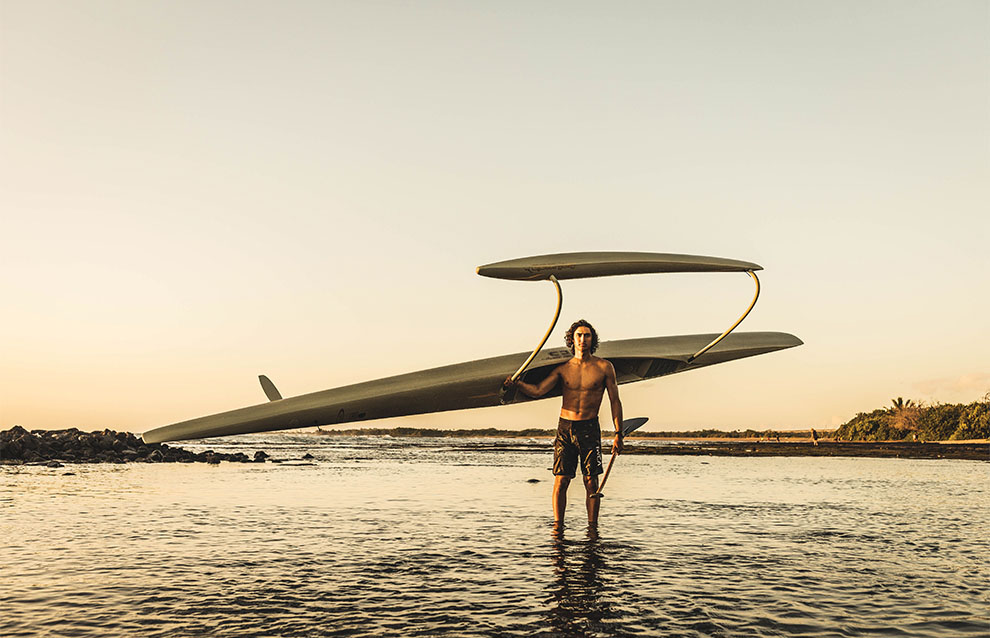 man holds boat on shoulder wades in water against sunset.