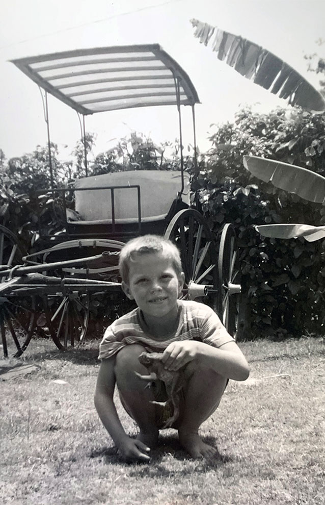 black and white photo of boy crouching outside next to a cart and large bushes.
