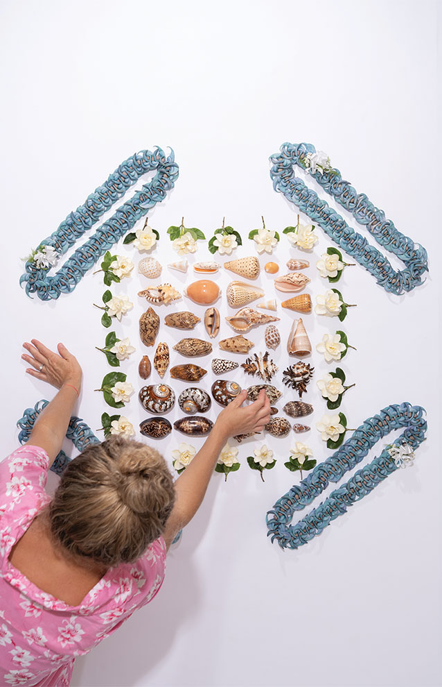 person in pink shirt arranges shells organized by colors of rainbow with blue leis arranged in symmetrical order.