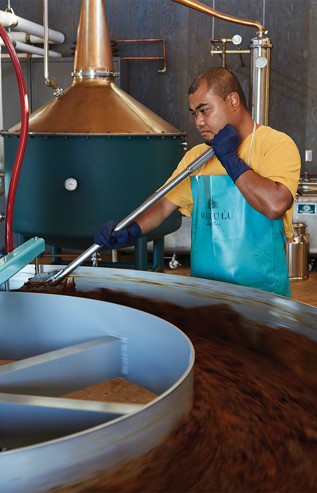 man in blue apron and black gloves stands in distillery operating a machine.