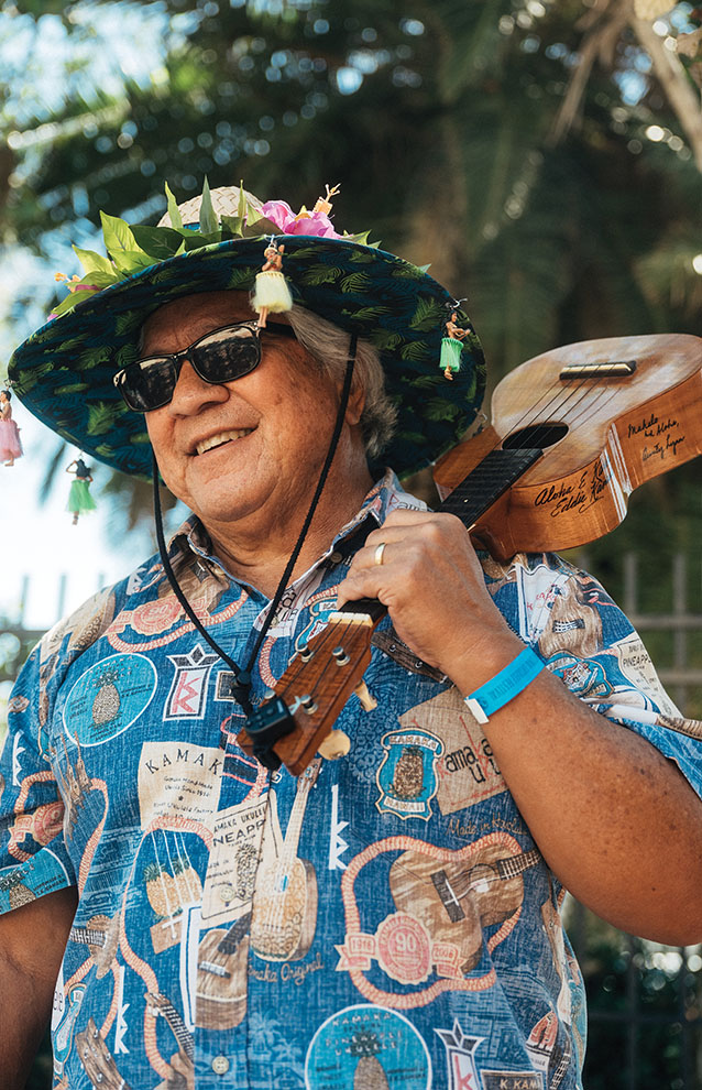 happy man in sunglasses and Hawaiian shirt holds string instrument over shoulder under a palm tree.