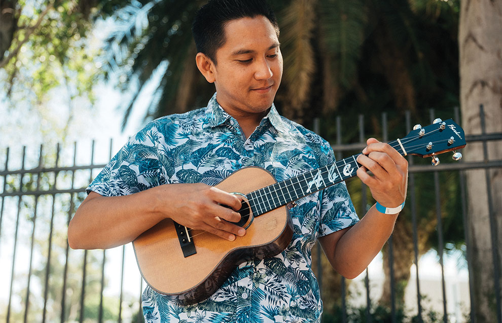 man in blue Hawaiian shirt plays a small string instrument by a fence under palm trees.