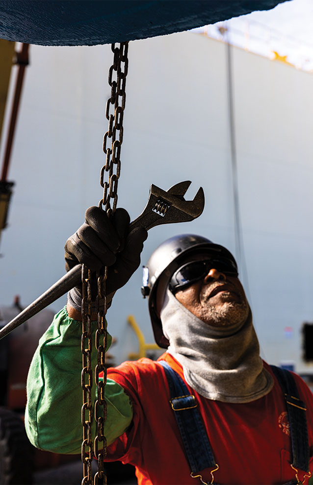 close up of construction worker holding onto a chain and a wrench dressed in helmet, vest, and glasses.