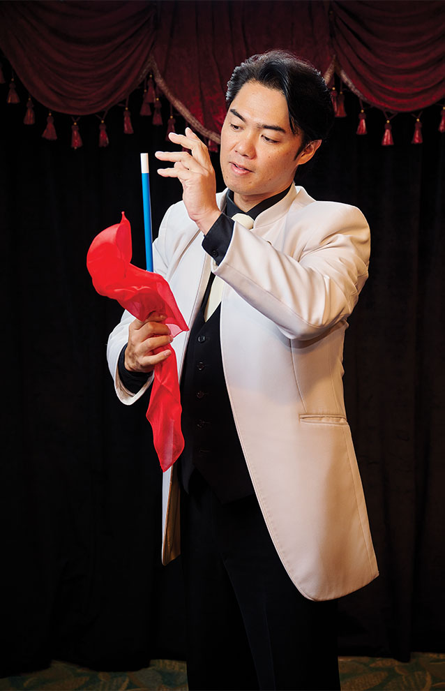 magician in white jacket holds magician's wand with a red tail of feathers attached.