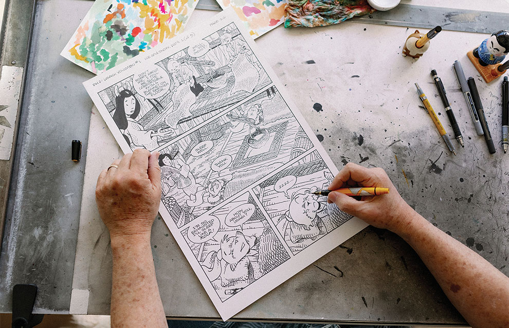 artist's hands sketch and hold a page of japanese anime on the artist's desk with pen and ink around.